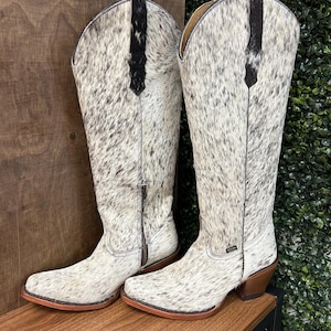 May include: A pair of tall, Western-style cowboy boots. The boots are primarily white with a speckled black pattern, featuring a brown sole and heel. A black leather accent runs down the top of the boot. The boots have a zipper closure.