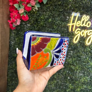 May include: A hand-painted ceramic napkin holder with a vibrant floral design in orange, red, yellow, and blue. The holder is rectangular with a blue border and a white interior. The background includes a neon sign that says "Hello gorg".