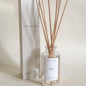 Könnte beinhalten: Ein klarer Glas-Reed-Diffusor mit Holzstäbchen und einem weißen Etikett mit der Aufschrift "SOHO" und "NORTH. ROSE". Eine weiße Schachtel mit dem Markennamen und Produktdetails steht hinter dem Diffusor. Der Diffusor enthält 118 ml Flüssigkeit.