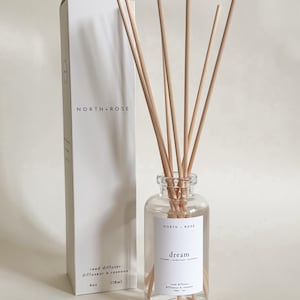 Peut inclure: Un diffuseur de roseaux en verre transparent avec des roseaux en bois et une étiquette blanche qui dit "dream". Le diffuseur est à côté d'une boîte blanche avec le texte "NORTH + ROSE". La boîte indique également "diffuseur de roseaux" et "118ml".