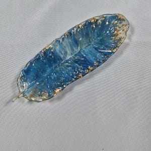 以下が含まれることがあります： 青と透明の樹脂で作られた、羽の形をした装飾トレイ。金箔のアクセントが施されています。トレイは詳細な羽のデザインで、長さは約20cmです。