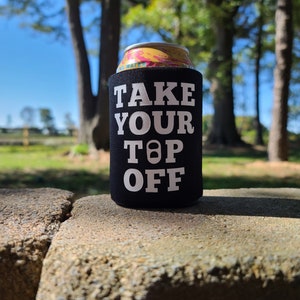 May include: Black and white can cooler with the text "TAKE YOUR TOP OFF" in white letters. The cooler is sitting on a gray stone surface.