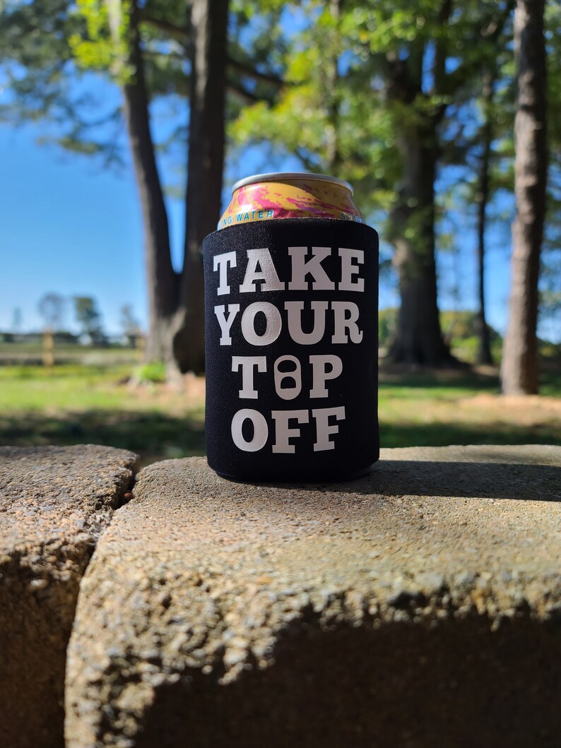May include: Black and white can cooler with the text "TAKE YOUR TOP OFF" in white letters. The cooler is sitting on a gray stone surface.