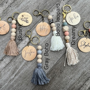 K&ouml;nnte beinhalten: Eine Sammlung von acht personalisierten Holz-Schl&uuml;sselanh&auml;ngern mit verschiedenen Perlen- und Quasten-Akzenten. Die Anh&auml;nger zeigen Namen, S&auml;tze wie "Just Breathe" und Muster wie "boho leopard".