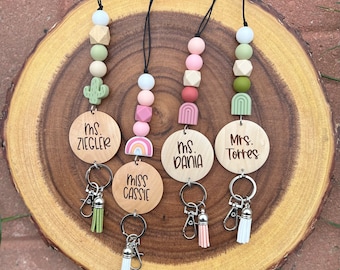 May include: Four wooden keychains with colorful beads and tassels. The keychains feature personalized name tags with the names "Miss Cassie", "Ms. Dania", "Ms. Ziegler", and "Mrs. Torres".