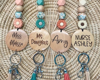 May include: Four personalized lanyards featuring wooden discs engraved with the names Miss Melissa, Ms. Douglas, Amy, and Nurse Ashley. Each lanyard includes a string of patterned, solid, and flower-shaped beads, a metal keyring, and a small tassel.
