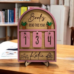 May include: A pink wooden book tracker with the words "Books Read This Year" and the name "Katie". It has three pink number cards showing "1", "3", and "4". The design includes small potted plants and floral details.