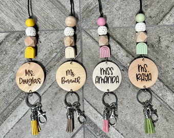 Puede incluir: Cuatro collares de llavero de madera con cuentas de colores y borlas. Los collares presentan etiquetas de nombre personalizadas con el texto "Ms. Douglas", "Ms. Bernath", "Miss Amanda" y "Ms. Raya".