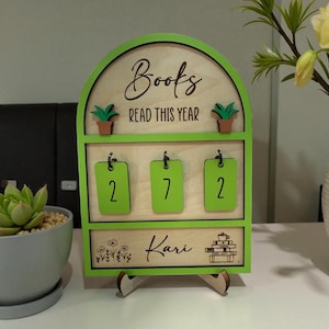May include: A wooden book tracker with a green frame. The top arch reads "Books READ THIS YEAR". Below are three green number tags displaying "2", "7", and "2". The bottom section is engraved with the name "Kari" and a stack of books with a teacup.