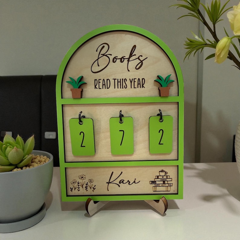 May include: A wooden book tracker with a green frame. The top arch reads "Books READ THIS YEAR". Below are three green number tags displaying "2", "7", and "2". The bottom section is engraved with the name "Kari" and a stack of books with a teacup.