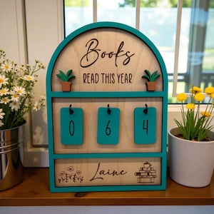 May include: Wooden book counter with teal accents. The sign reads "Books READ THIS YEAR" with the numbers "064" displayed. The name "Laine" is also featured. Two small potted plants are on top.