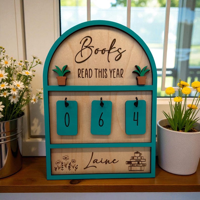 May include: Wooden book counter with teal accents. The sign reads "Books READ THIS YEAR" with the numbers "064" displayed. The name "Laine" is also featured. Two small potted plants are on top.