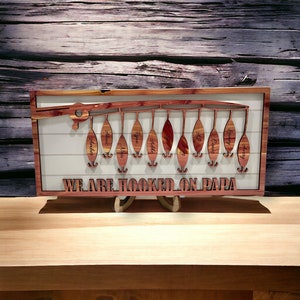 Op de afbeelding: Een houten wanddecoratie in de vorm van een hengel met de tekst "We are hooked on Papa" en 11 haken met namen op elke haak. De namen zijn Amber, Alexis, Lila, Ryan, Addy, T, AJ, Caiden, Casey, Avery.