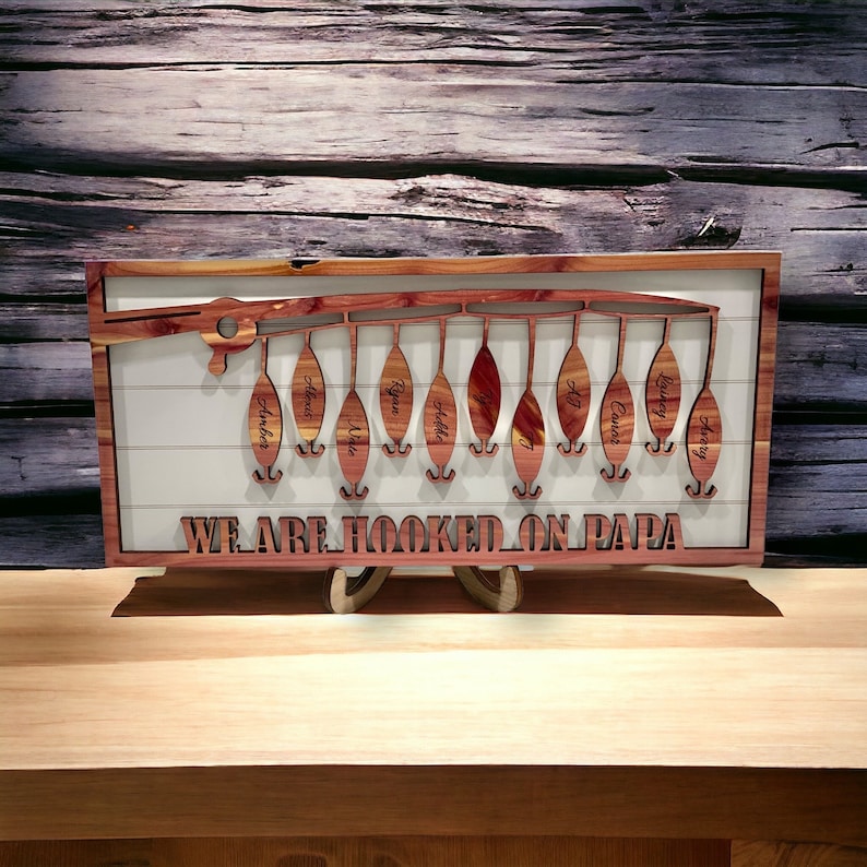 Op de afbeelding: Een houten wanddecoratie in de vorm van een hengel met de tekst "We are hooked on Papa" en 11 haken met namen op elke haak. De namen zijn Amber, Alexis, Lila, Ryan, Addy, T, AJ, Caiden, Casey, Avery.