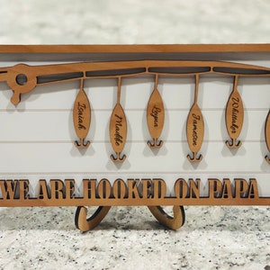 Op de afbeelding: Een houten bord met een hengel en vijf haken, elk met een naamschildje. Het bord staat "We are hooked on Papa".