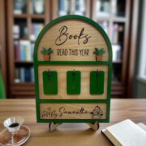 May include: A wooden book tracker with a green frame. The top section reads "Books READ THIS YEAR" and has two small potted plants. Below are three green key holders. The bottom section is personalized with the name "Samantha" and a stack of books.