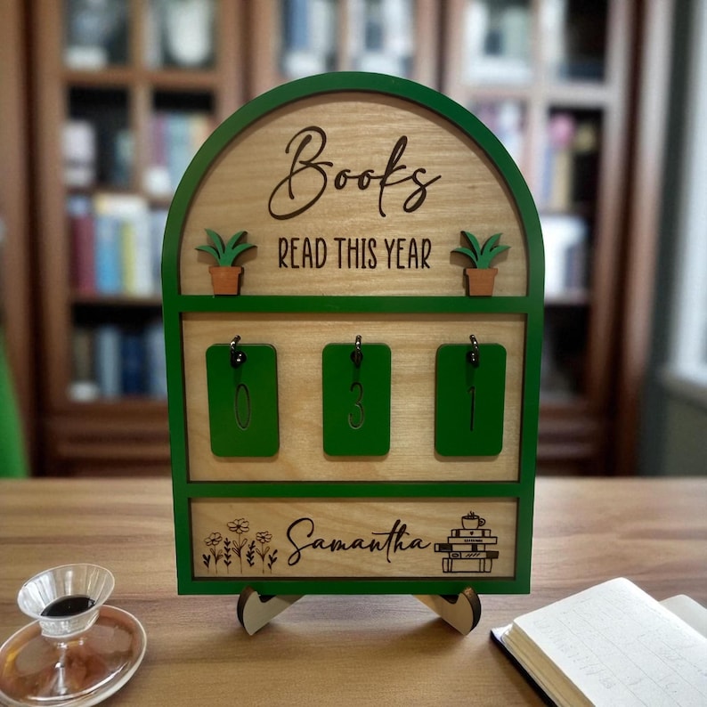 May include: A wooden book tracker with a green frame. The top section reads "Books READ THIS YEAR" and has two small potted plants. Below are three green key holders. The bottom section is personalized with the name "Samantha" and a stack of books.