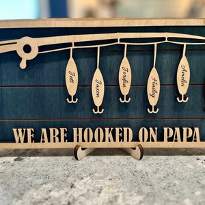 May include: A wooden sign with a blue background and a cutout of a fishing rod with hooks. The hooks have names on them: Jett, Jaxon, Jordan, Hailey, Amelia, and Brynlee. The sign reads "We are hooked on Papa".