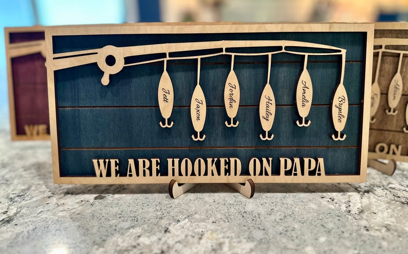 May include: A wooden sign with a blue background and a cutout of a fishing rod with hooks. The hooks have names on them: Jett, Jaxon, Jordan, Hailey, Amelia, and Brynlee. The sign reads "We are hooked on Papa".