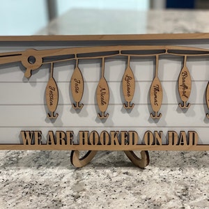 Op de afbeelding: Een houten bord met een ontwerp van een hengel en zes hengels die eraan hangen. Elke hengel heeft een naam: Jessica, Josie, Nicale, Luciana, Thomas, Bradford en Makayla. Het bord staat "We are hooked on Dad".