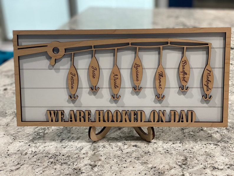 Op de afbeelding: Een houten bord met een ontwerp van een hengel en zes hengels die eraan hangen. Elke hengel heeft een naam: Jessica, Josie, Nicale, Luciana, Thomas, Bradford en Makayla. Het bord staat "We are hooked on Dad".