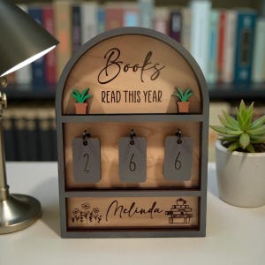 May include: Wooden book counter with the words "Books Read This Year" and the number "266" displayed on hanging tags. The counter has a gray frame and includes small potted plants and the name "Melinda".