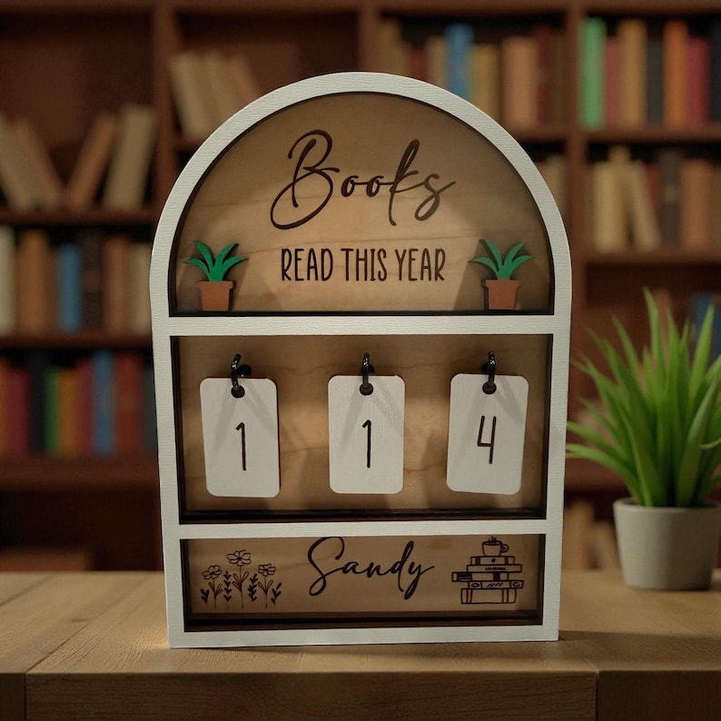 May include: A white wooden book counter with the words "Books READ THIS YEAR" and the number "114" displayed. The counter has a wooden background and includes small potted plants and the name "Sandy".