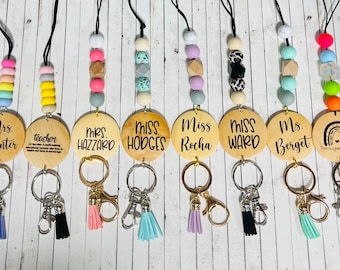 May include: Ten wooden keychains with colorful beads and tassels. Each keychain has a different name on it, including "Mrs. Hunter", "Teacher", "Mrs. Hazzard", "Miss Hodges", "Miss Rocha", "Miss Ward", "Ms. Berget", and "Teach, Love, Inspire".
