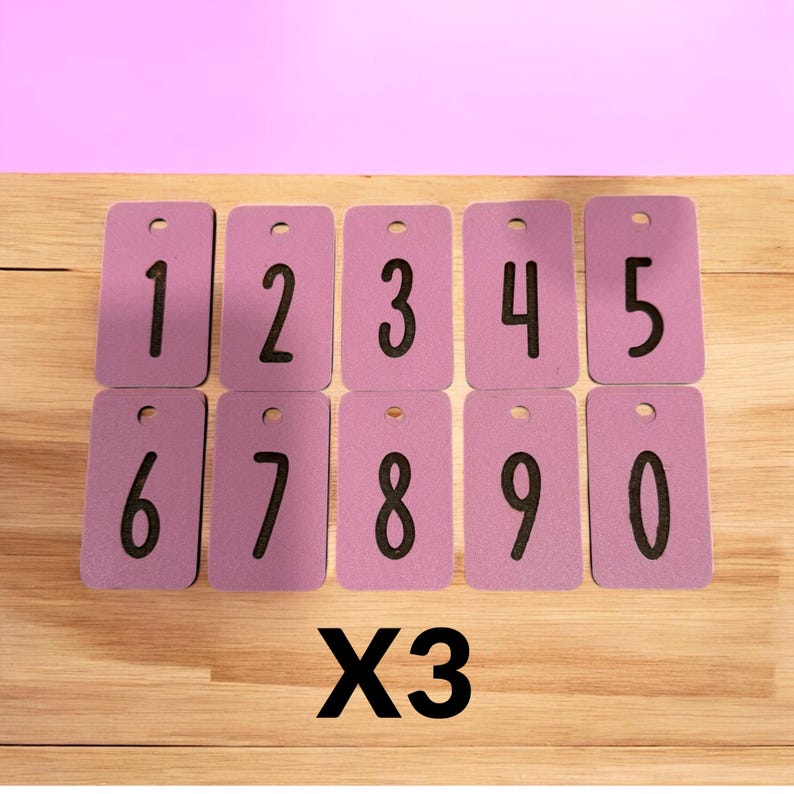 May include: A set of ten lavender number tags, each with a black number from 0 to 9. The tags are rectangular with a small hole at the top. The text "X3" is below the tags. The background is a light wood surface.
