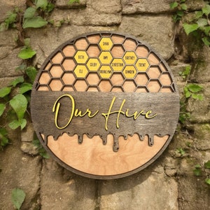 May include: A round wooden sign with a honeycomb design and the words "Our Hive" in yellow script. The honeycomb is filled with yellow names, including Dana, Beth, Amy, Colby, Eli, Brooklyn, Christian, Camden, Britney, and Trent.