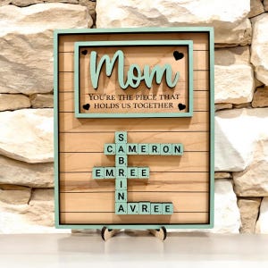 May include: Wooden sign with a light green frame and accents. The sign reads "Mom" and "YOU'RE THE PIECE THAT HOLDS US TOGETHER". Below, names are spelled out using Scrabble tiles. The sign is displayed on a wooden stand.