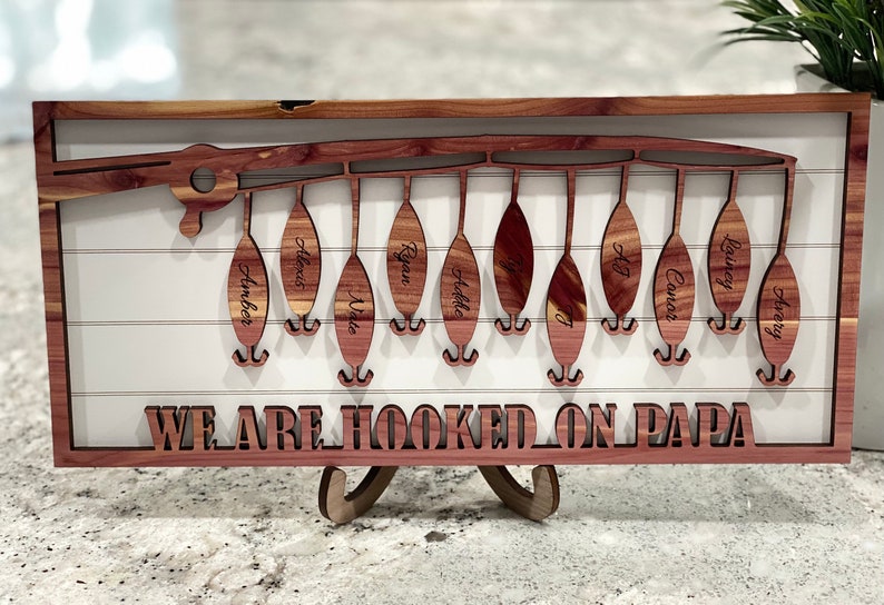 Op de afbeelding: Een houten wanddecoratie met een ontwerp van een hengel en gepersonaliseerde namen op elke lok. De tekst "WE ARE HOOKED ON PAPA" staat onder de lokken.