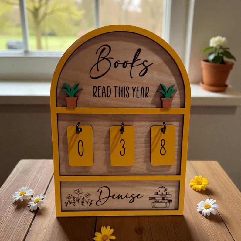 May include: A yellow wooden book counter with the text "Books READ THIS YEAR" and the numbers "038". The counter has a wooden background and includes the name "Denise" with a floral and book design. Two small potted plants are on top.