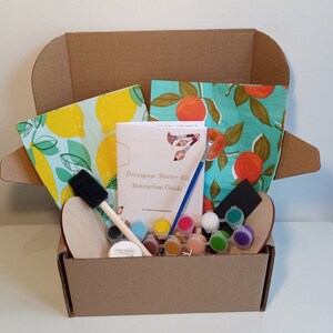 May include: A cardboard box filled with a decoupage starter kit. The kit includes a wooden craft blank, paintbrushes, paint, and two patterned fabric squares. The box also includes an instruction guide titled "Decoupage Starter Kit Instruction Guide".