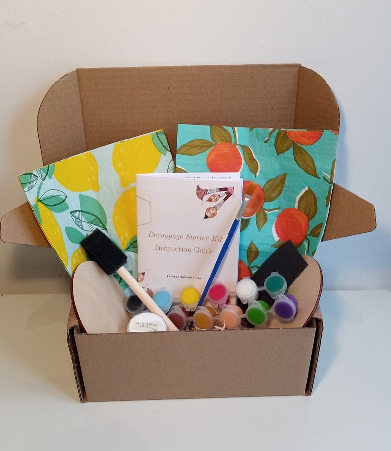May include: A cardboard box filled with a decoupage starter kit. The kit includes a wooden craft blank, paintbrushes, paint, and two patterned fabric squares. The box also includes an instruction guide titled "Decoupage Starter Kit Instruction Guide".