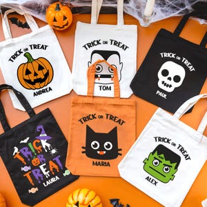 Op de afbeelding: Zes witte en zwarte Halloween trick-or-treat tassen met verschillende Halloween-figuren en namen erop gedrukt. De tassen liggen op een oranje oppervlak met een spinnenweb en pompoenen.