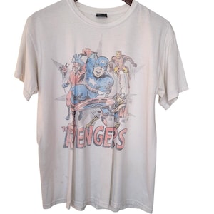 May include: A white short-sleeve t-shirt featuring a faded graphic print of The Avengers comic book characters, including Captain America, Iron Man, and Thor, with the text The Avengers printed in red and black lettering on the front.