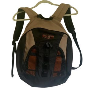May include: A tan, black, and brown Eddie Bauer backpack with adjustable black shoulder straps. The backpack has a black mesh pocket and a red and black Eddie Bauer logo. The backpack is hanging on a white hanger.