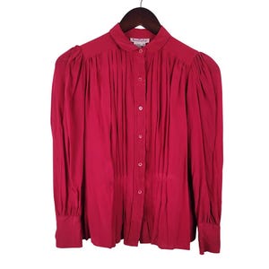 May include: A long-sleeved button-down shirt in a deep magenta color. The shirt features a small collar, vertical pleats running down the front, and gathered shoulders. It has a buttoned front closure and cuffs, and a label reading Amelie.