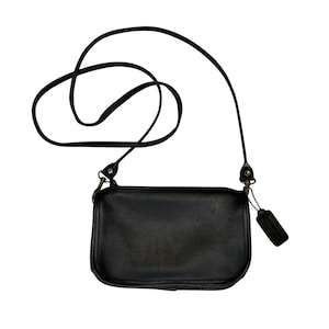 Könnte beinhalten: Schwarze Leder-Umhängetasche mit einem langen, dünnen Riemen. Die rechteckige Tasche hat abgerundete Ecken und ein kleines, angebrachtes Etikett. Der Riemen ist verstellbar, und die Tasche scheint Vintage zu sein.