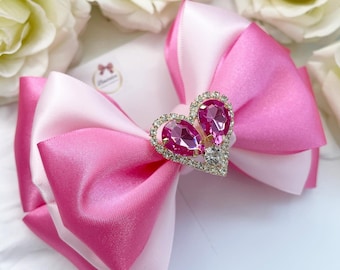 Puede incluir: Un lazo de satén rosa y blanco con un adorno de corazón de diamantes de imitación dorados. El lazo es perfecto para añadir un toque de elegancia a cualquier atuendo.