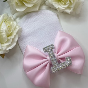 May include: White baby hat with a pink satin bow and a rhinestone letter L.