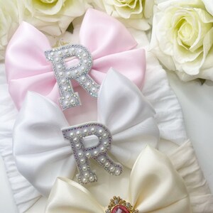 May include: Three satin hair bows with pearl embellishments. The top bow is pink, the middle bow is white, and the bottom bow is cream. The pink and white bows have a pearl letter "R" in the centre. The cream bow has a pink rhinestone in the centre.