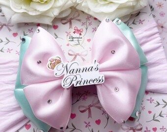 Puede incluir: Un lazo para el pelo de capas en cintas de satén rosa claro y verde menta, adornado con pedrería transparente. Una placa central blanca muestra un gráfico de corona dorada y el texto negro Nanna's Princess sobre una diadema rosa suave.