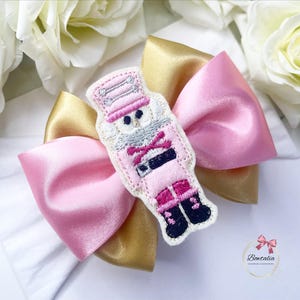 May include: A pink and gold satin bow with a felt nutcracker appliqué. The nutcracker is wearing a pink and white uniform with a black belt and a red sash. The bow is ideal for adding a festive touch to any outfit.