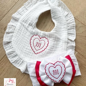 May include: A white muslin baby bib with a ruffled edge and a pink embroidered heart featuring the letter m, paired with a matching white headband featuring a large white and pink bow with a central heart patch embroidered with the letter m.