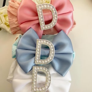 May include: Three satin hair bows with pearl-encrusted letter D charms. The bows are pink, blue, and white.
