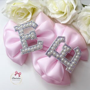 May include: Two pink satin bows with the letter "E" in white with pearl and rhinestone embellishments.