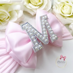 May include: A pink satin bow with a white letter M embellished with pearls and rhinestones. The letter M is centred on the bow.
