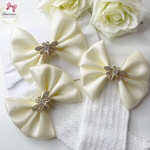 May include: Three ivory satin bows with gold bee embellishments. The bows are attached to white knitted socks.